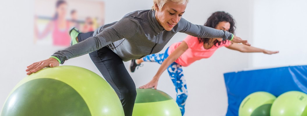 Fitte kaukasische Frauen machen eine Kranich-Übung auf einem Bein stehend, wobei sie ihre Arme seitlich ausstrecken und sich an einem Balance-Ball festhalten, während sie im Fitness-Club trainieren